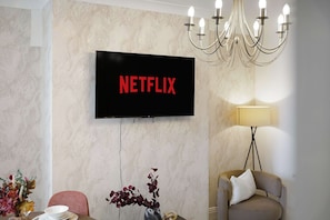 Ein Zimmer mit montiertem Fernseher, der 'NETFLIX' in roten Buchstaben zeigt, einer Kandelaber, einem Tisch mit Blumenvase und einem Sofa.