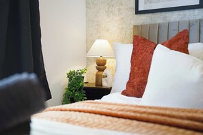 Ein ordentlich bezogenes Bett mit einer weiß-orangenen Decke, ein Nachttisch mit einer Lampe und eine kleine grüne Pflanze.