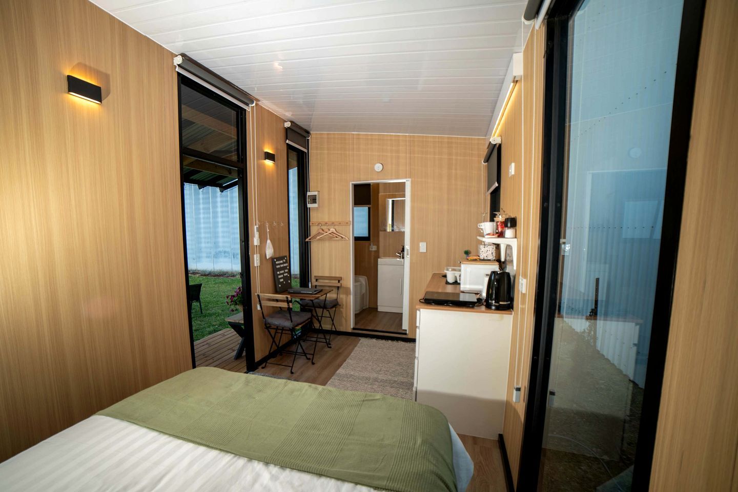 A compact hotel room with a bed, a small kitchenette, a dining area, and a sliding glass door leading to a balcony.