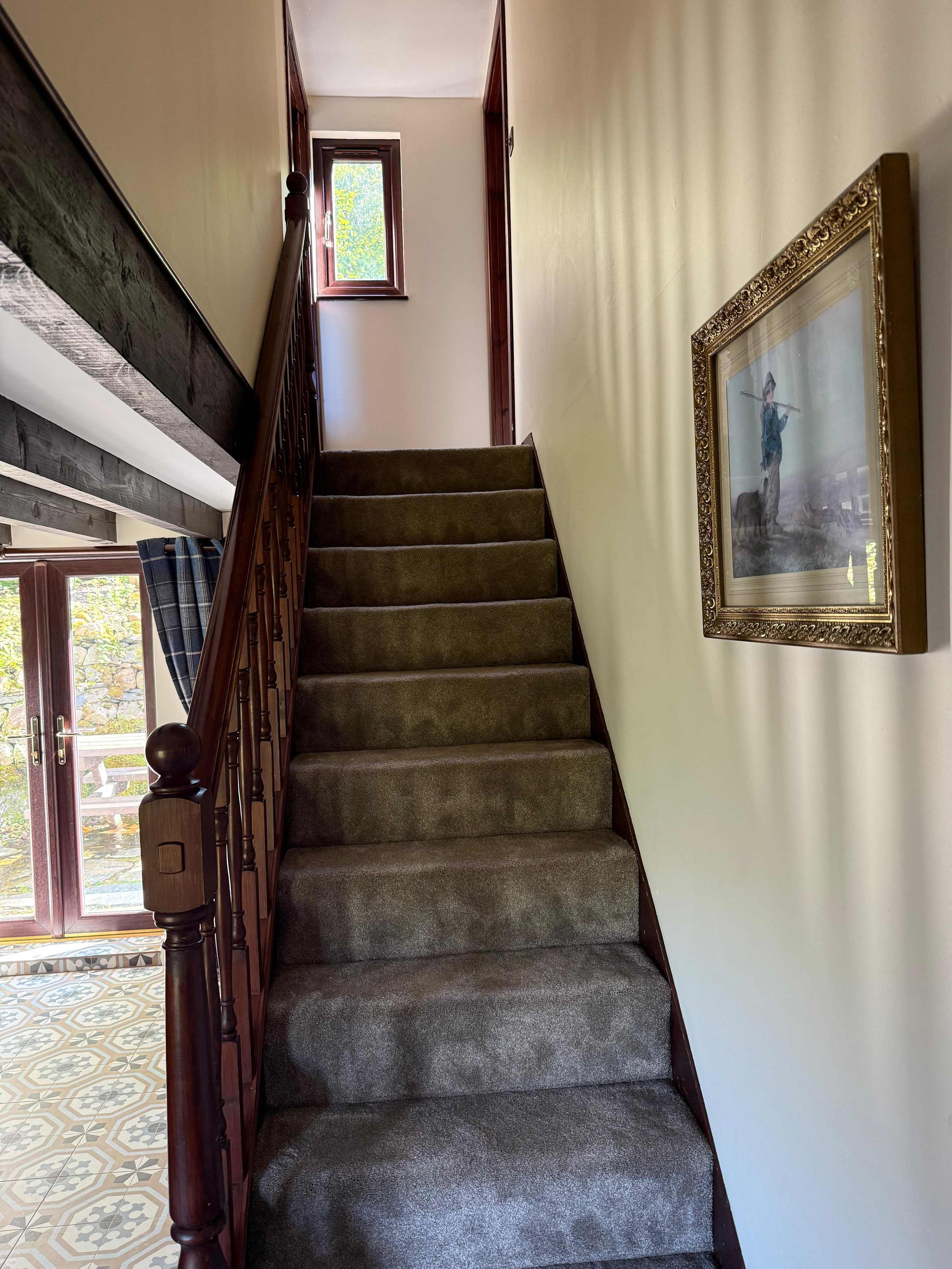 Un escalier avec des marches recouvertes de moquette et des balustrades en bois, menant à un palier doté d’une petite fenêtre.