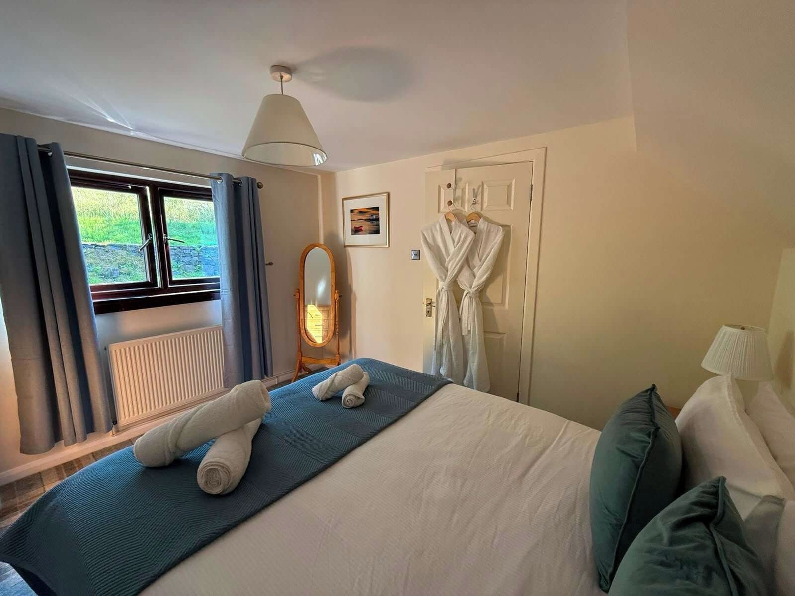 A hotel room with a bed, a window with curtains, a radiator, a mirror, and a robe hanging on the wall.