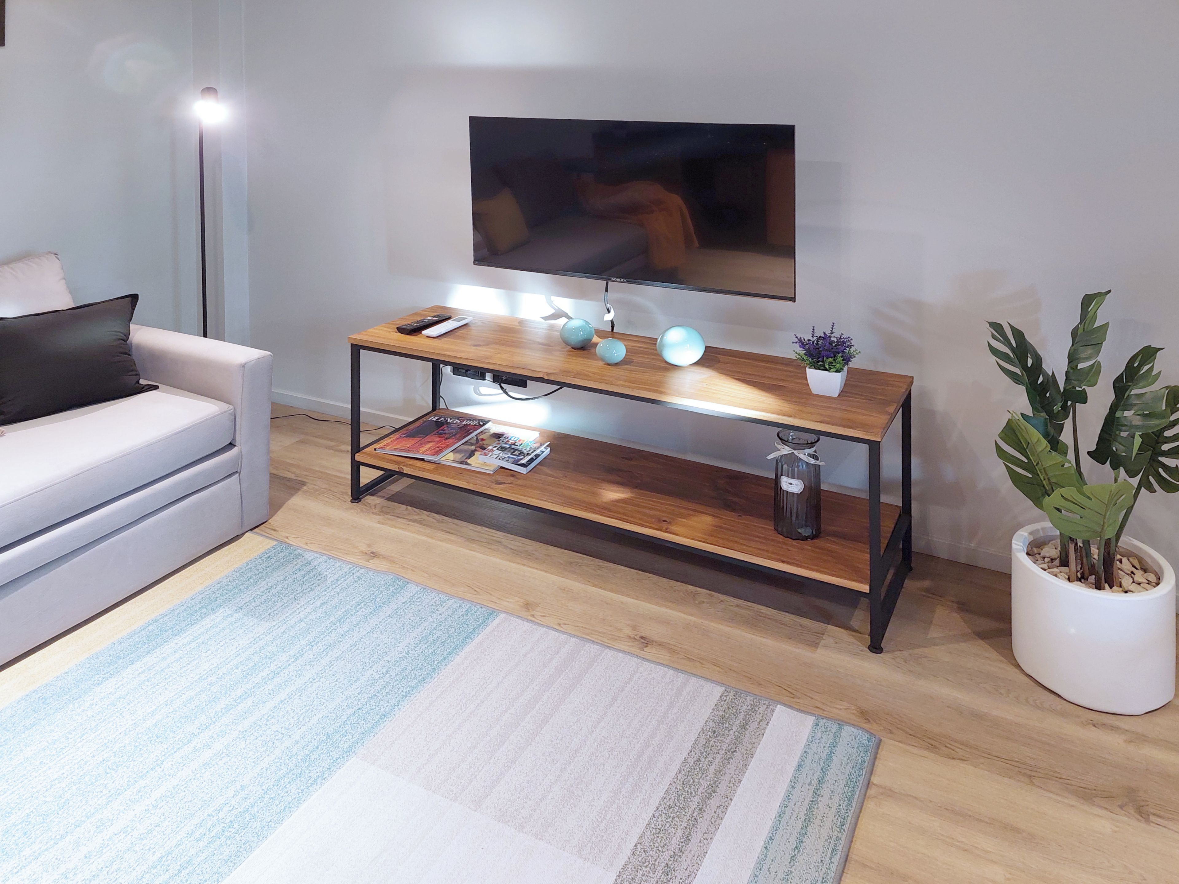 Una sala moderna con un sofá, una mesa de centro de madera con objetos decorativos, un televisor montado en la pared y una planta en maceta.