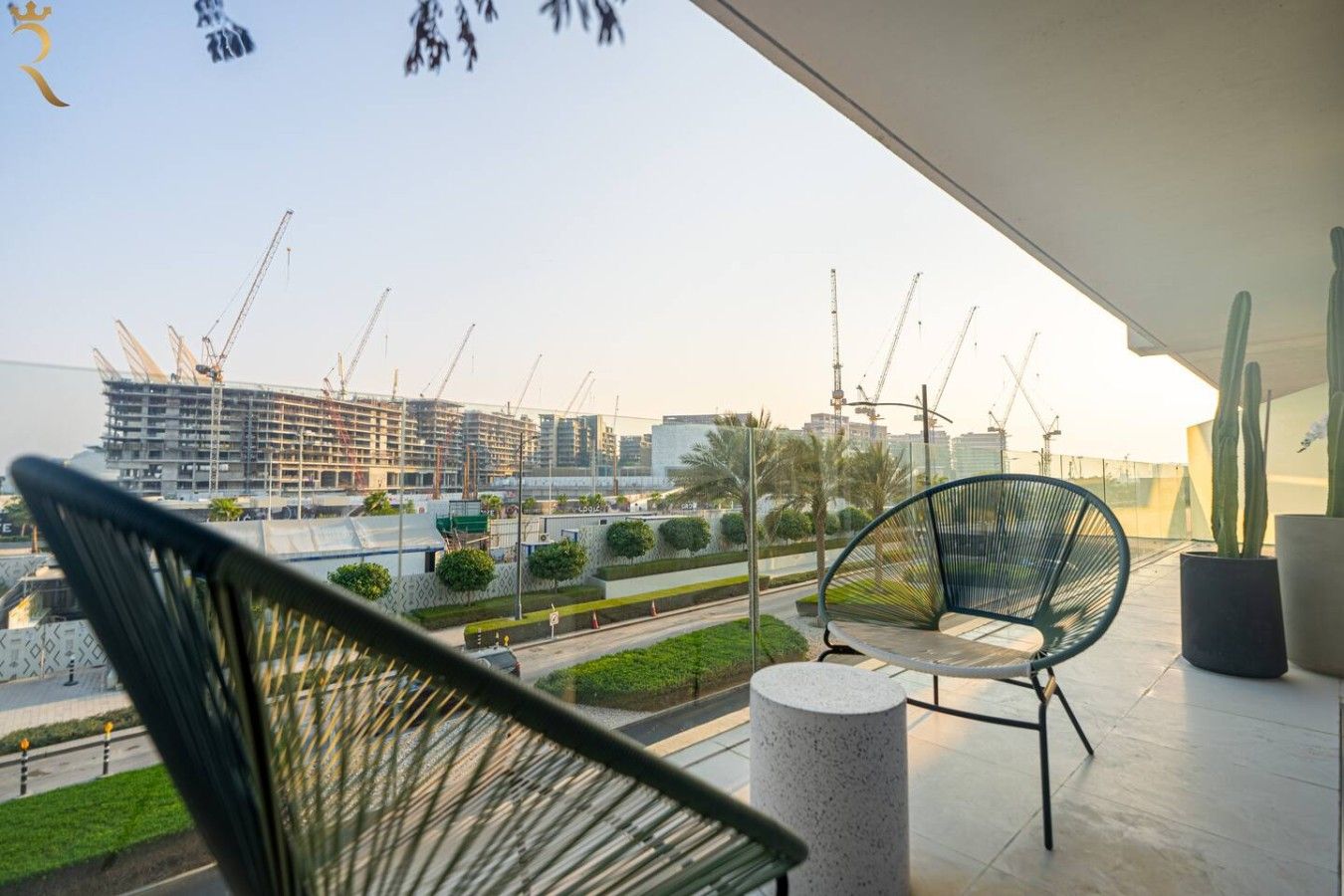 A balcony with two chairs, a cactus, and a view of a construction site with cranes.