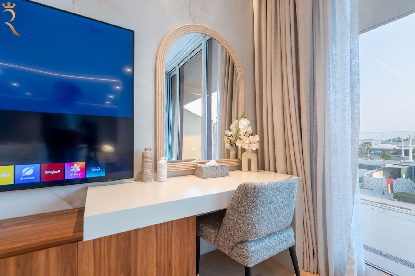 A hotel room with a large flat-screen TV, a mirror, a vanity with a mirror, and a window with curtains.