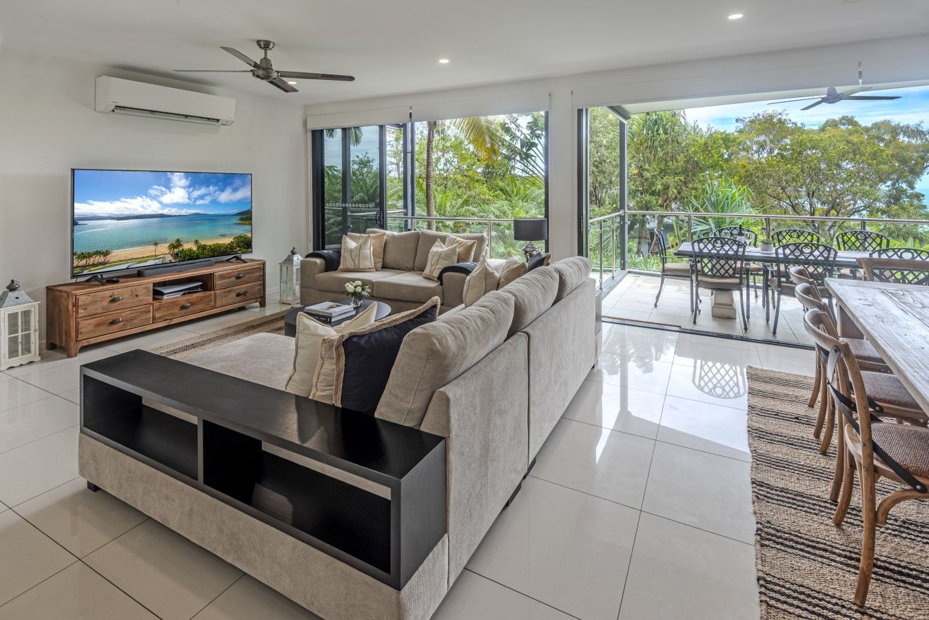 A modern living room with a large flat-screen TV, a sectional sofa, and a dining area with outdoor seating.