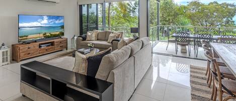 A modern living room with a large flat-screen TV, a sectional sofa, and a dining area with outdoor seating.