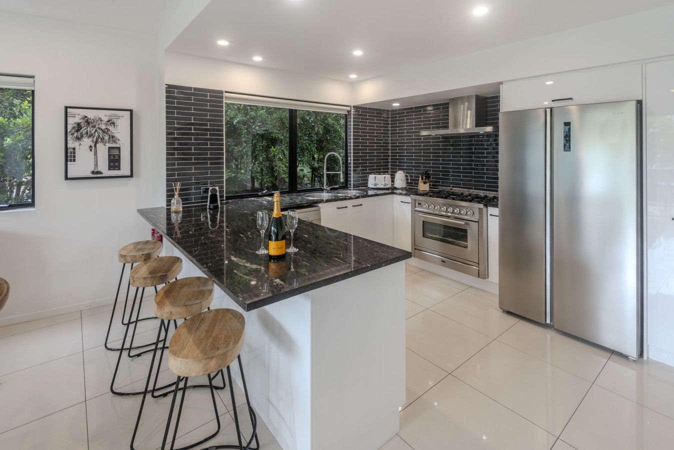 A modern kitchen with a central island, bar stools, a stainless steel refrigerator, and a large window.