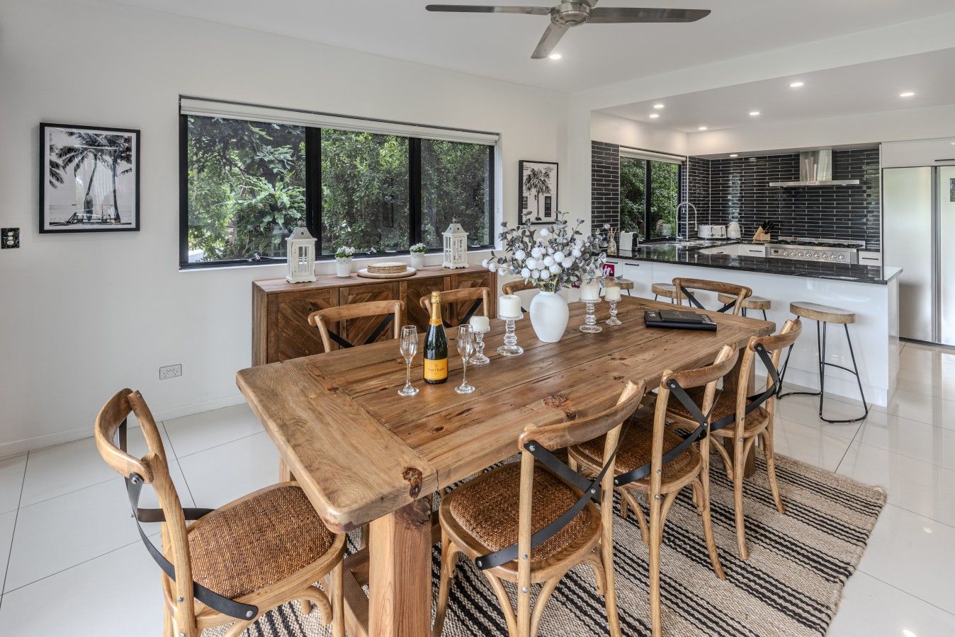 A modern dining area with a wooden table and chairs, a kitchen in the background, and large windows offering a view of greenery.