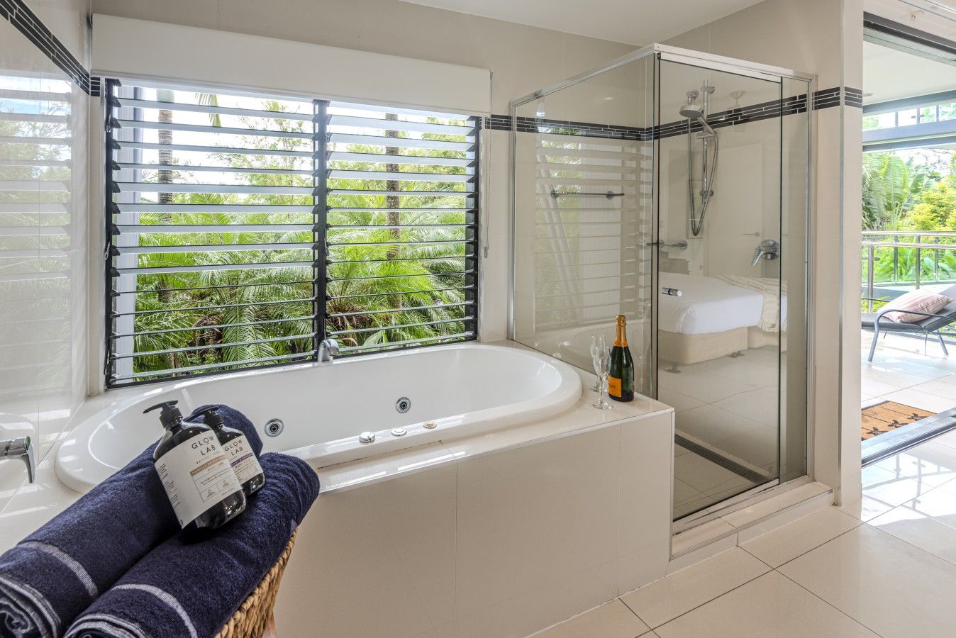 Une salle de bain moderne dotée d’une douche à parois de verre, d’une grande baignoire et d’une fenêtre avec stores donnant sur un espace verdoyant.