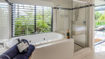 Une salle de bain moderne dotée d’une douche à parois de verre, d’une grande baignoire et d’une fenêtre avec stores donnant sur un espace verdoyant.