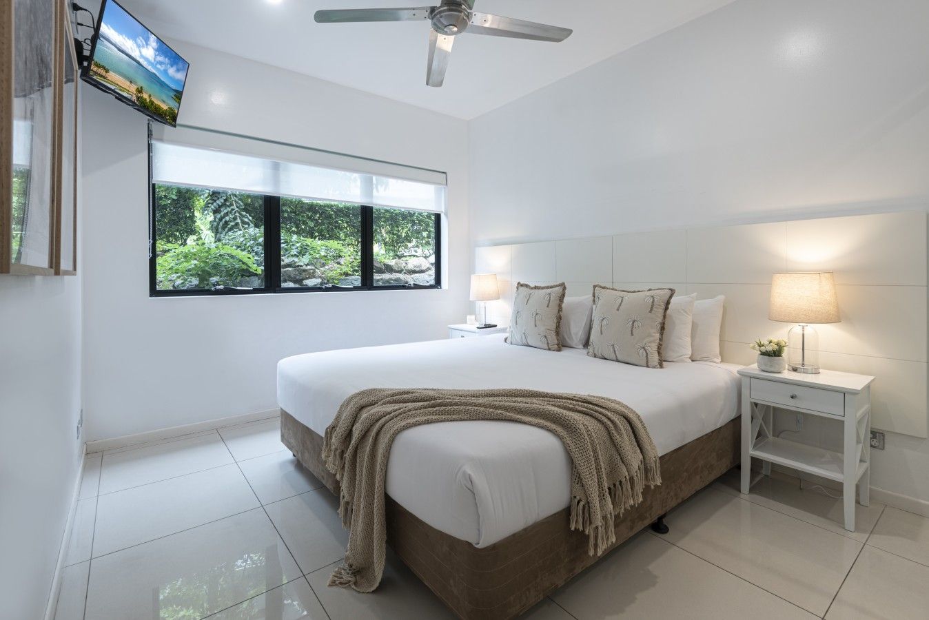 A bedroom with a large bed, a television mounted on the wall, a ceiling fan, and a window with a view of greenery.
