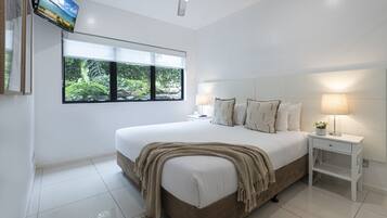 A bedroom with a large bed, a television mounted on the wall, a ceiling fan, and a window with a view of greenery.