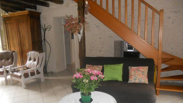 Uma sala de estar com escada de madeira, um sofá com almofadas, uma cadeira e uma mesa com planta em vaso.