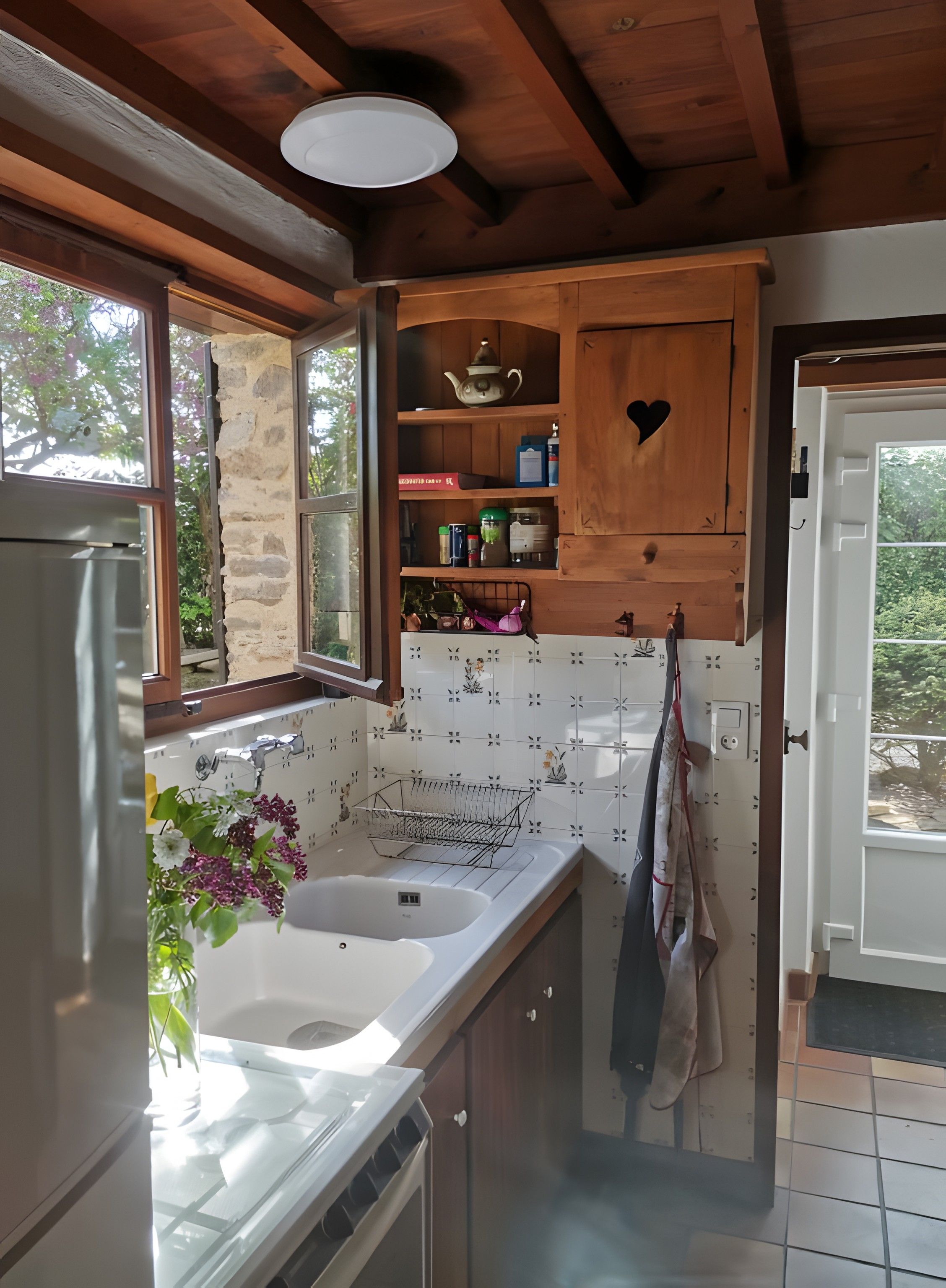 Une cuisine avec un mur en pierre, des armoires en bois, un évier blanc et une fenêtre donnant sur un paysage verdoyant.