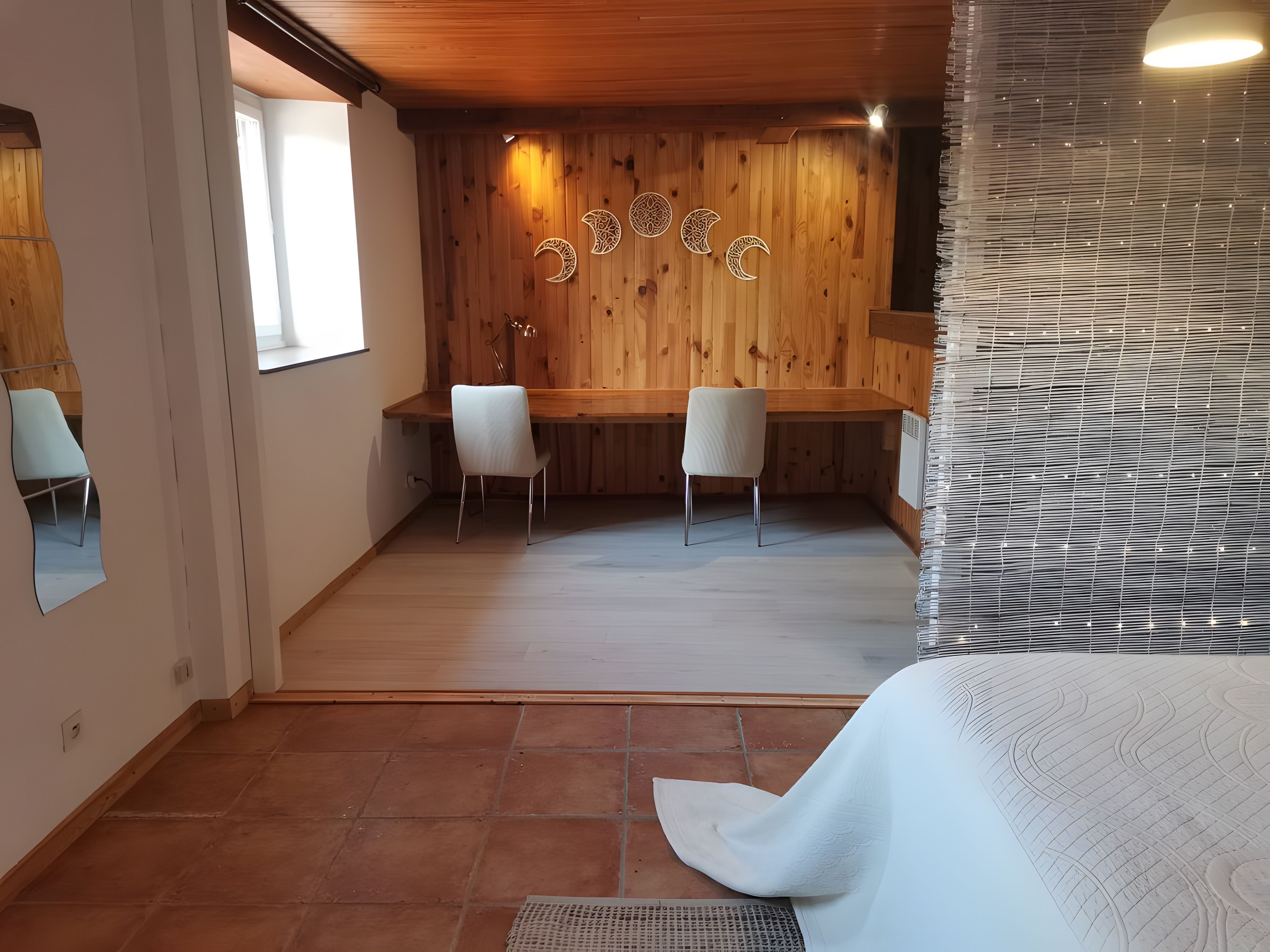 A room with a wooden ceiling, a desk with two chairs, a large window, and a wall with decorative moon shapes.