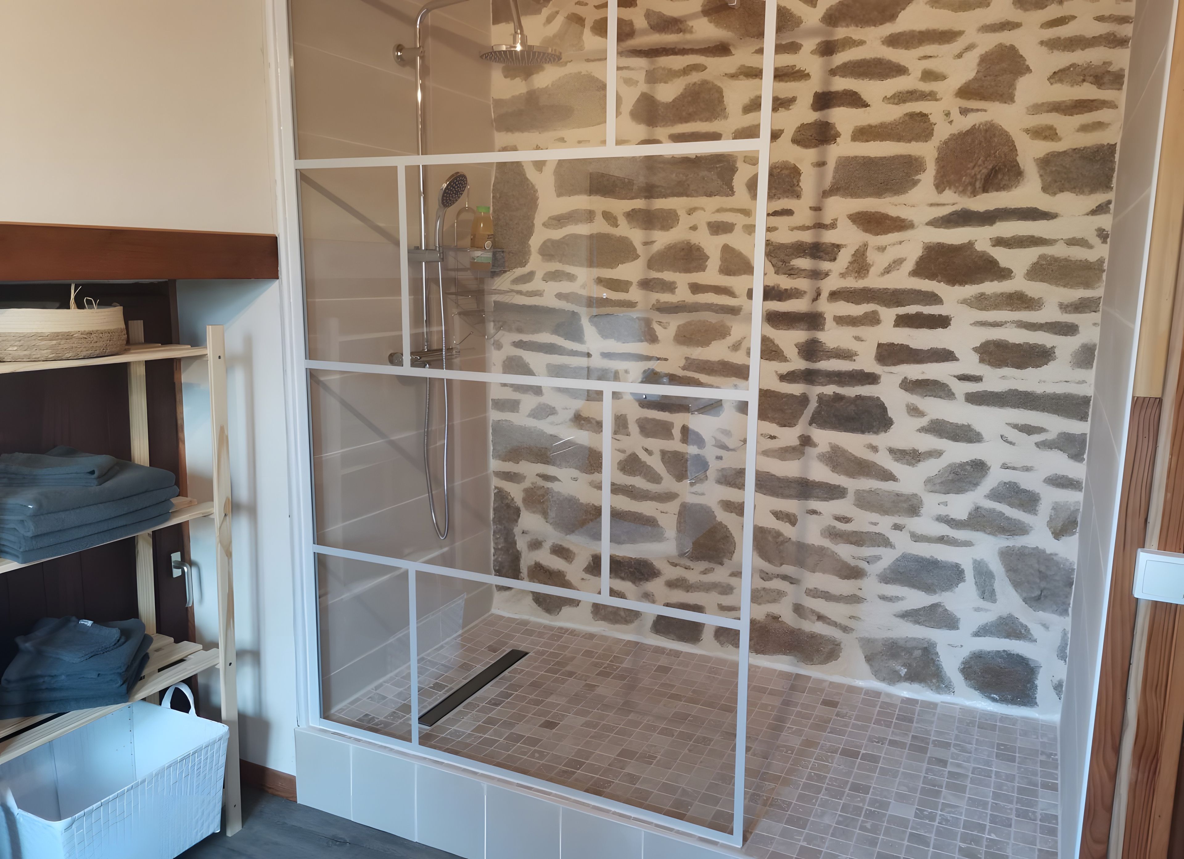 A shower area with a stone wall, a glass door, and a shelf with folded towels.
