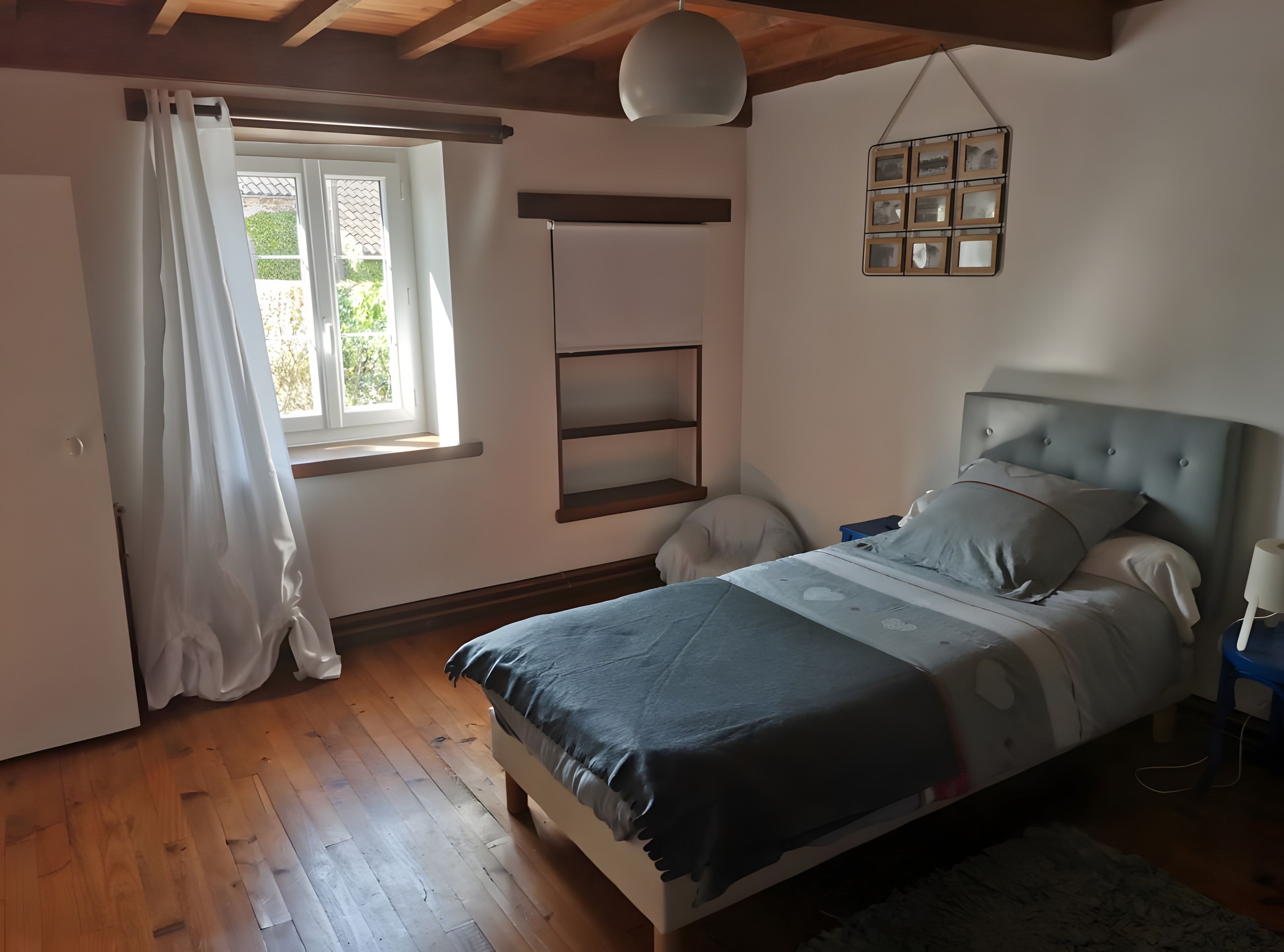 Une chambre à coucher avec un lit, une fenêtre avec des rideaux, une table de chevet et une étagère fixée au mur.