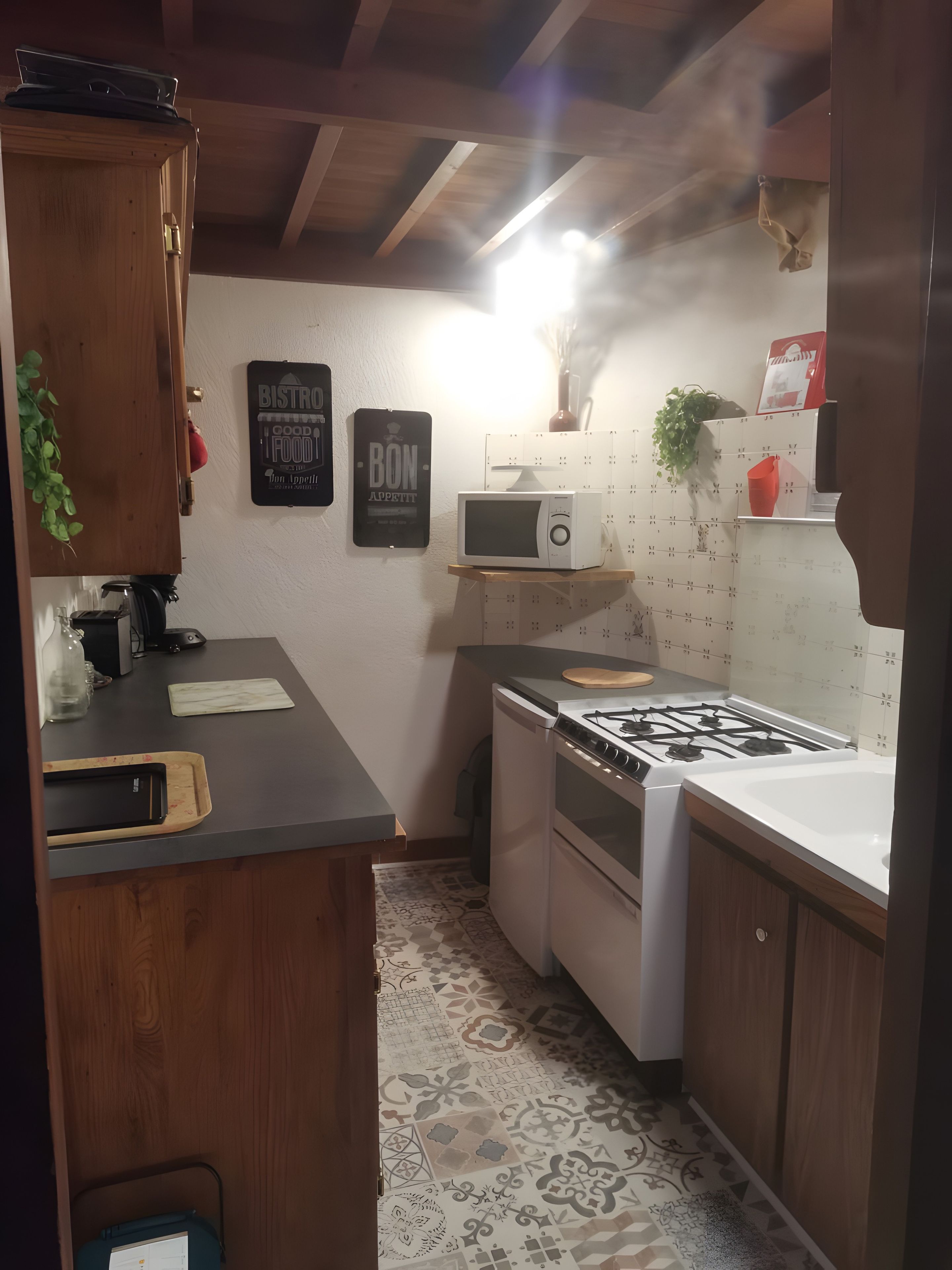 Cocina compacta con microondas, estufa y refrigerador pequeño. Se observan armarios de madera y piso con diseño.