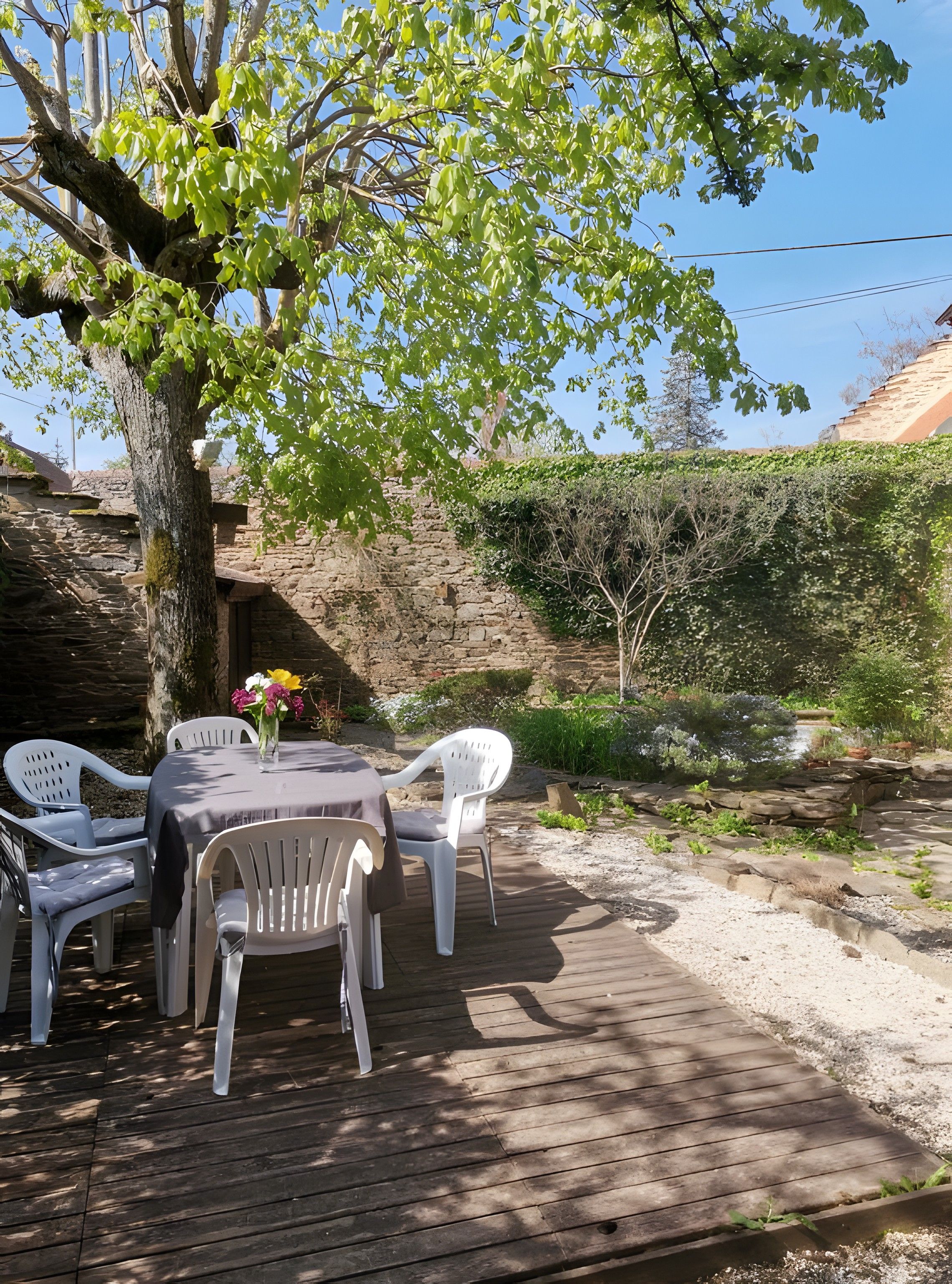A wooden deck with a table and chairs, surrounded by greenery and a stone wall.