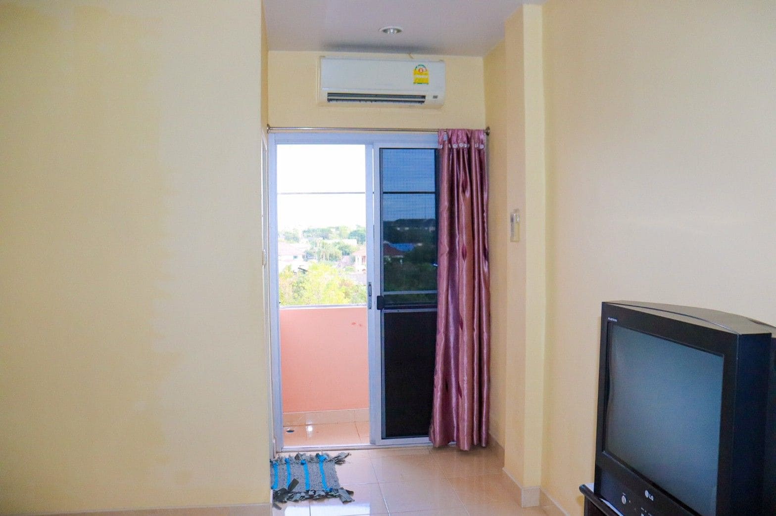 A room with a television, an air conditioning unit, a sliding glass door leading to an outdoor area, and a tiled floor.