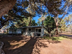 A small, single-story cabin with a porch, surrounded by trees and a clear sky.