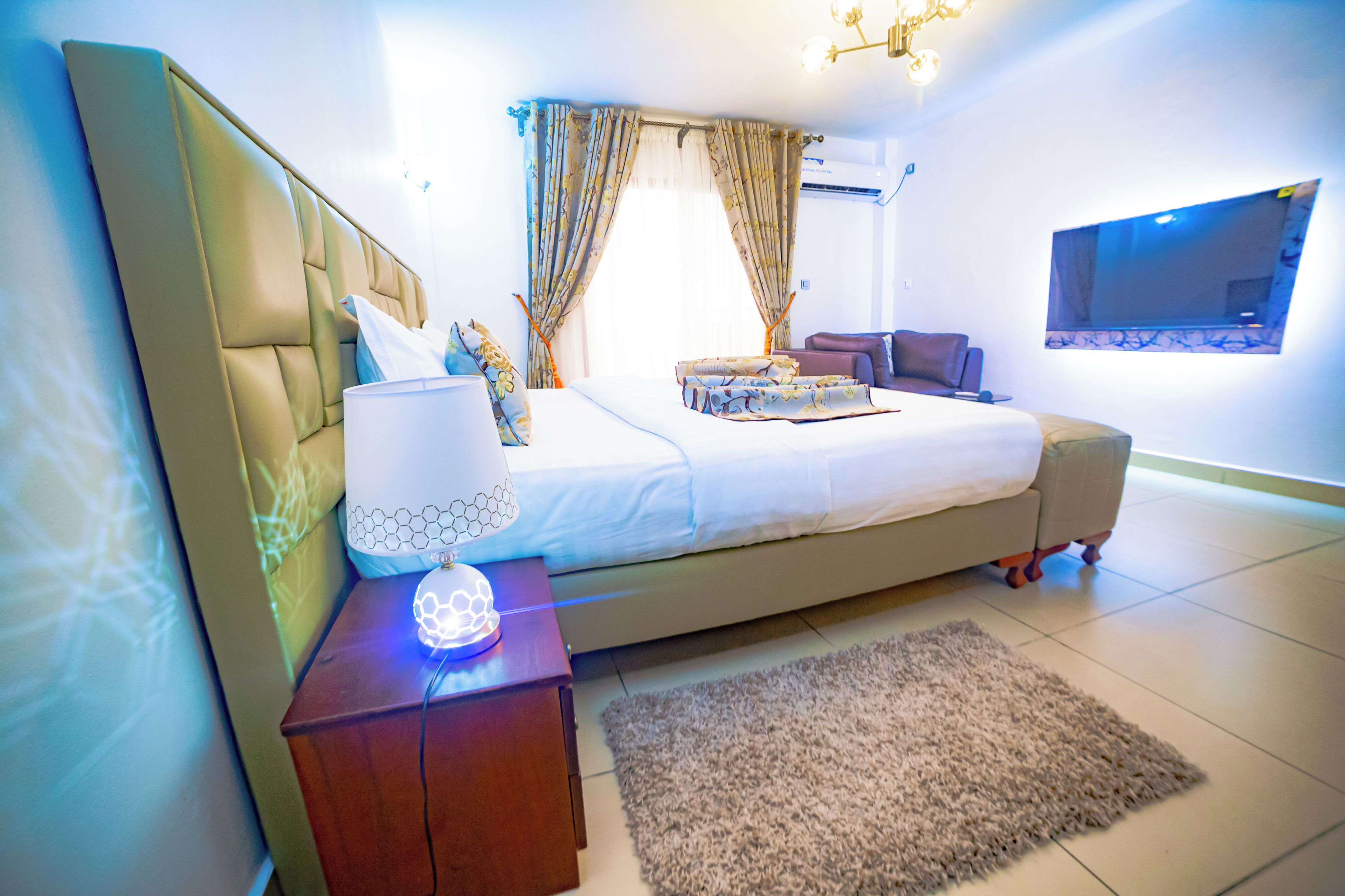 Een moderne slaapkamer met een groot bed, een nachtkastje met een lamp, een aan de muur bevestigde televisie en een raam met gordijnen.
