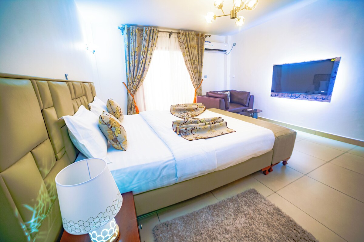 Habitación de hotel con una cama grande, una mesita de noche con lámpara, un sofá y un televisor en la pared.