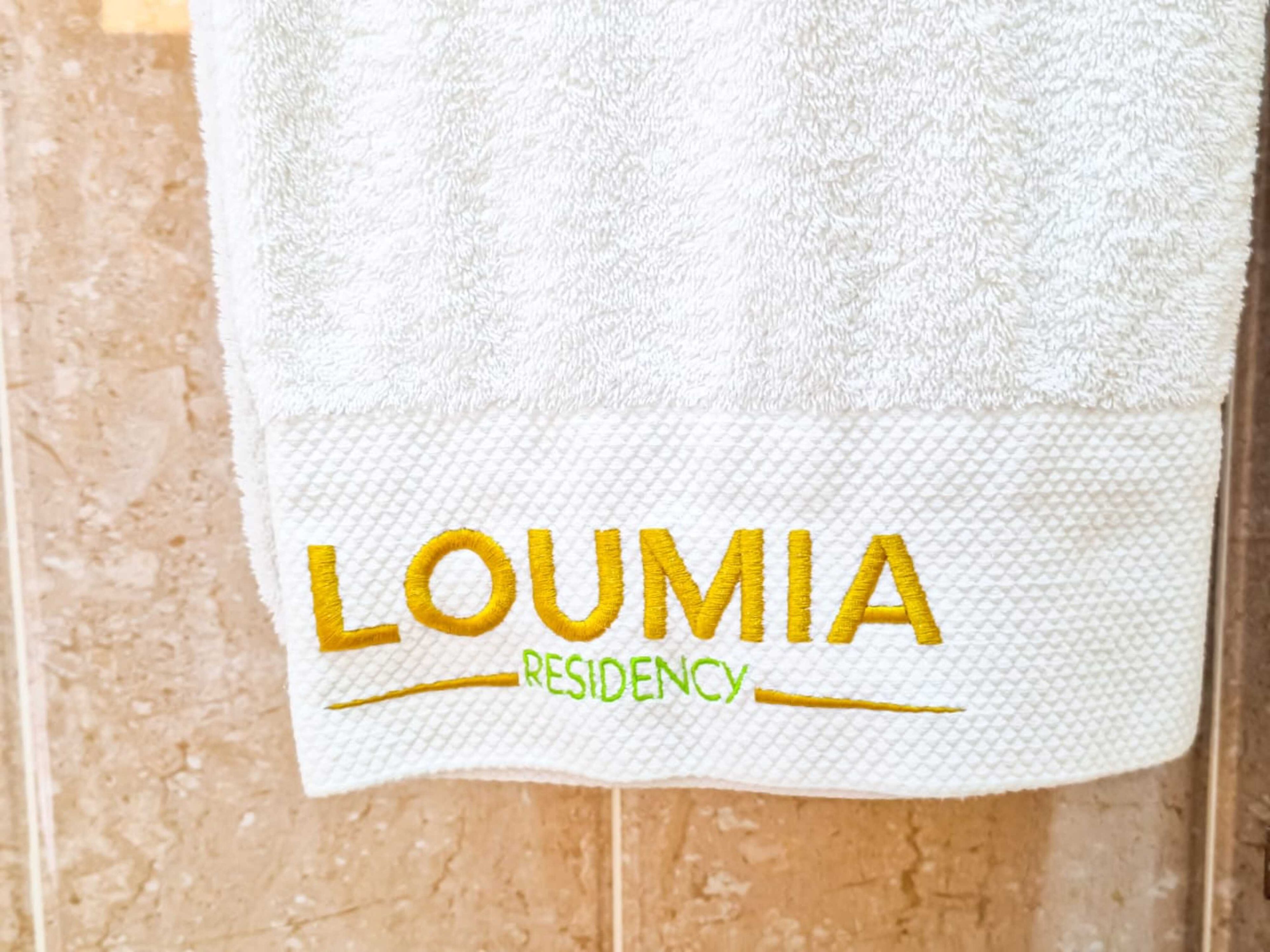 A white towel with embroidered text 'LOUMIA RESIDENCY' on a textured surface.