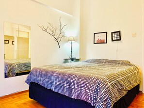 Een slaapkamer met een bed, een nachtkastje, een lamp, een spiegel en wanddecoratie.