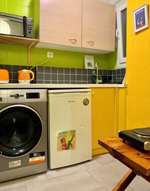 A compact kitchen with a washing machine, a small refrigerator, and a microwave. - Athens Home (Athina)