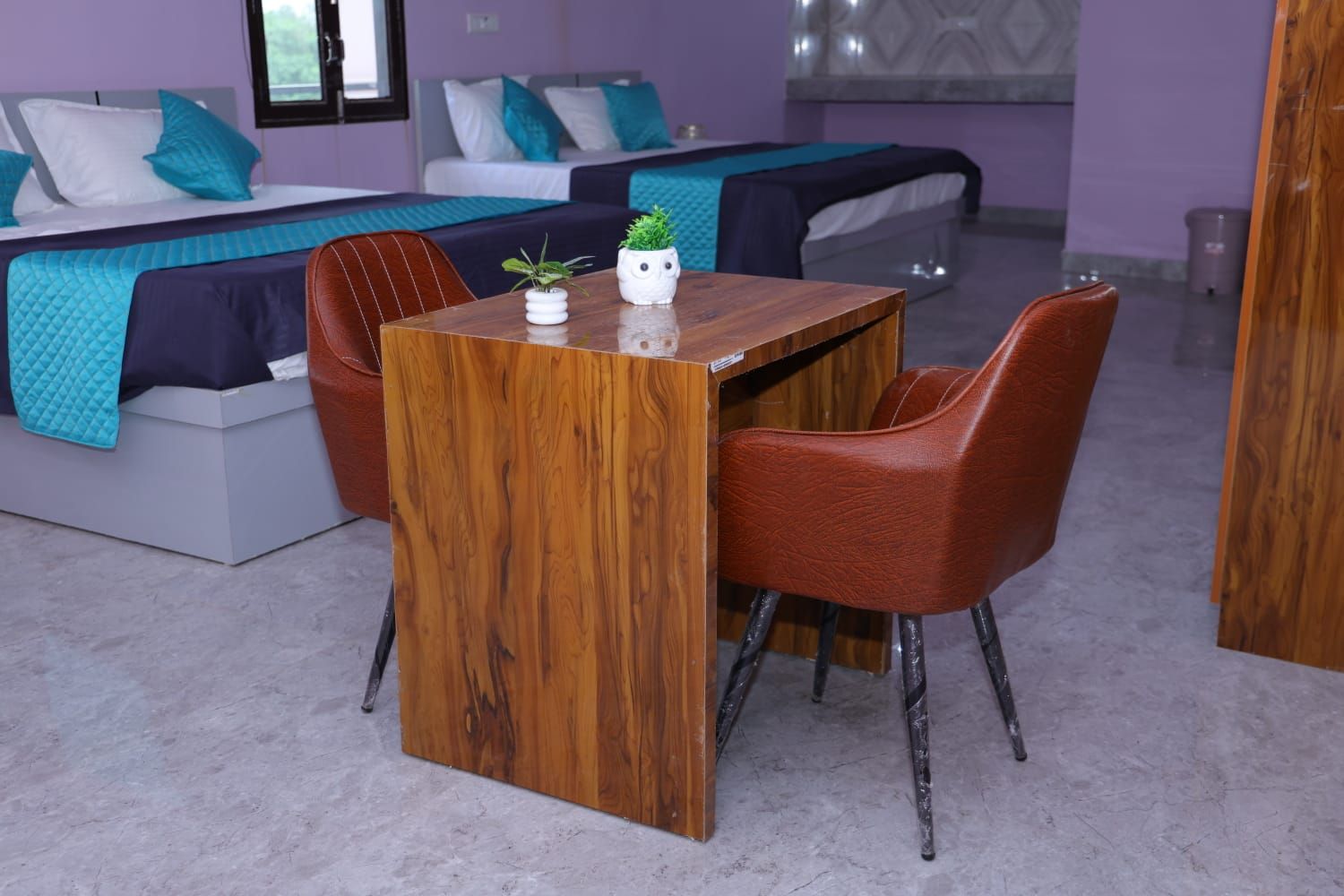 A modern hotel room with a wooden desk, two chairs, a small table with a plant, and a bed with blue and white bedding.
