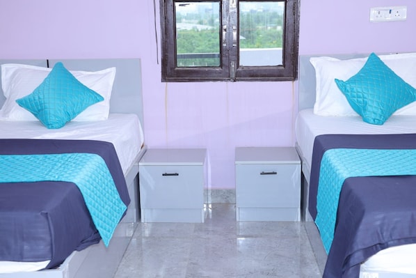 A hotel room with two beds, each with a blue quilted bedspread and a matching pillow. There are two bedside tables with drawers, and a window above the beds. - THE ROYAL KING (Gurugram)