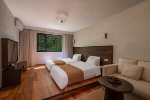 Habitación de hotel con dos camas, un sofá, una mesita y un ventanal con vistas a árboles.