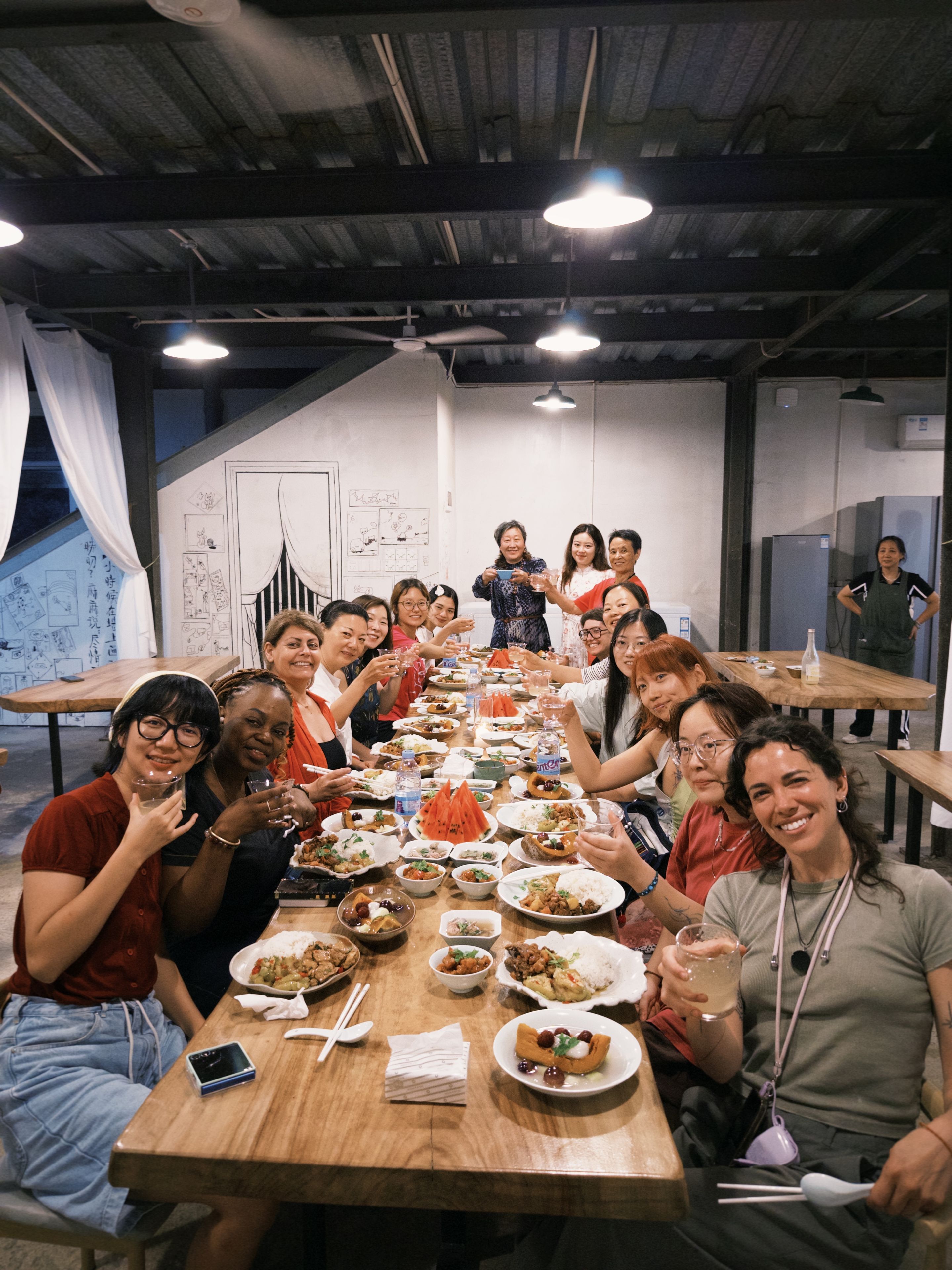 A group of people seated around a long wooden table with various dishes, in a room with exposed ceiling beams and a whiteboard on the wall.