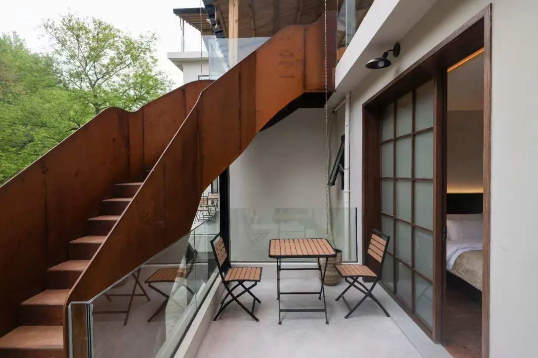 Un interior moderno con una escalera, barandilla de cristal y una zona de estar con mesas y sillas.