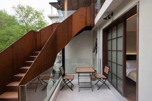 Un interior moderno con una escalera, barandilla de cristal y una zona de estar con mesas y sillas.