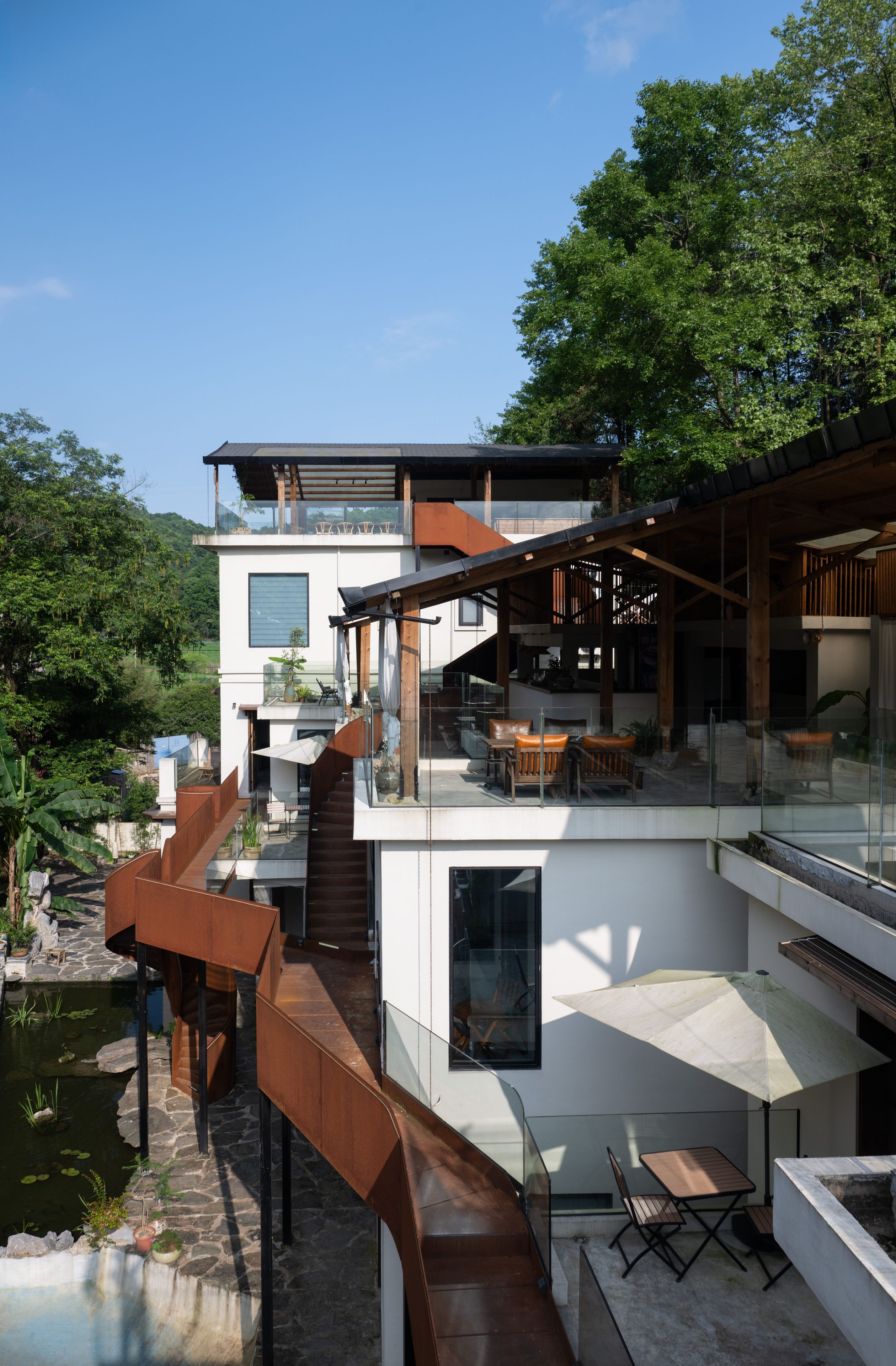 A modern multi-level house with a glass balcony, outdoor seating, and a small pond.