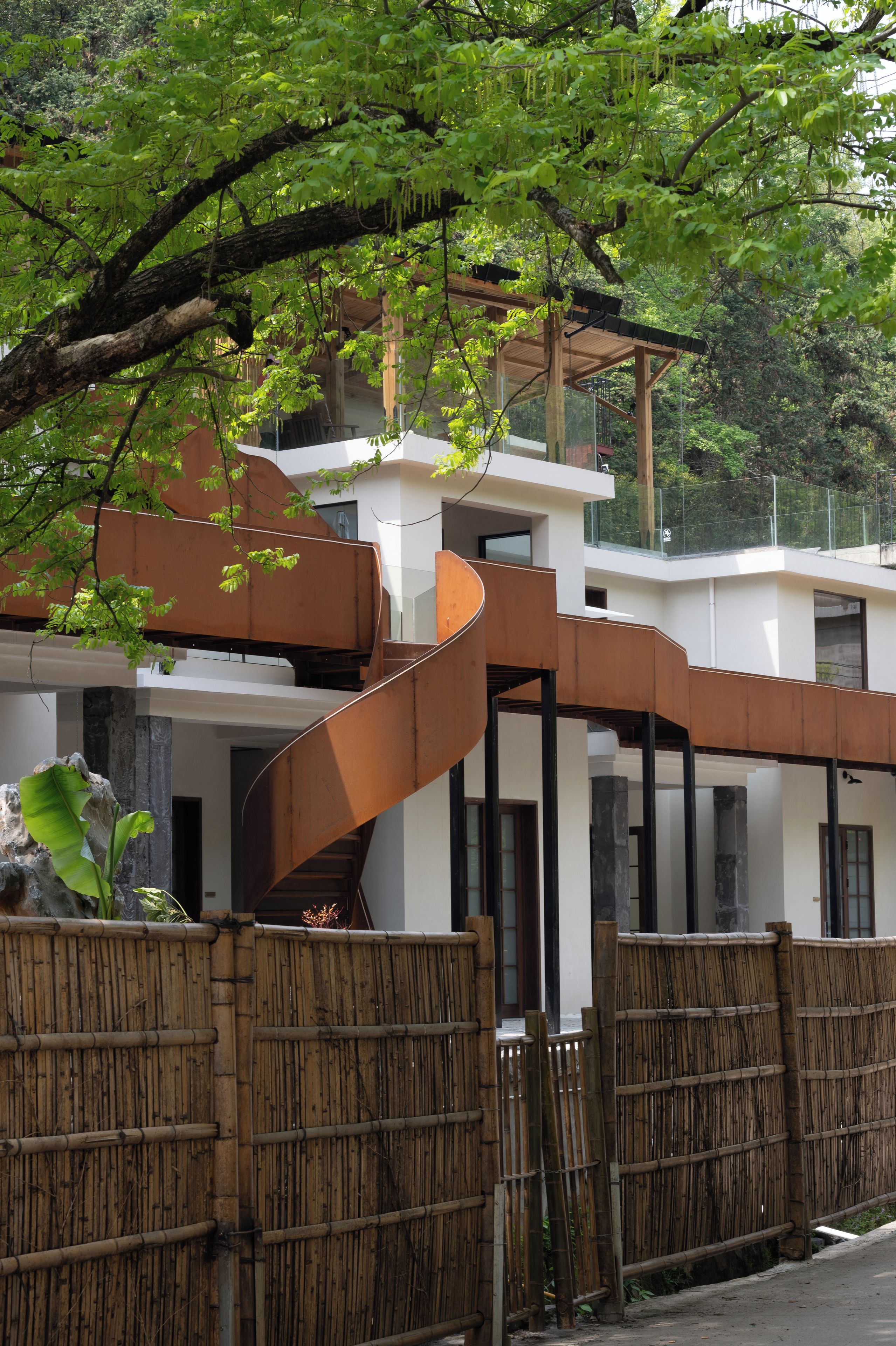 A modern building with a spiral staircase, wooden fence, and lush greenery.