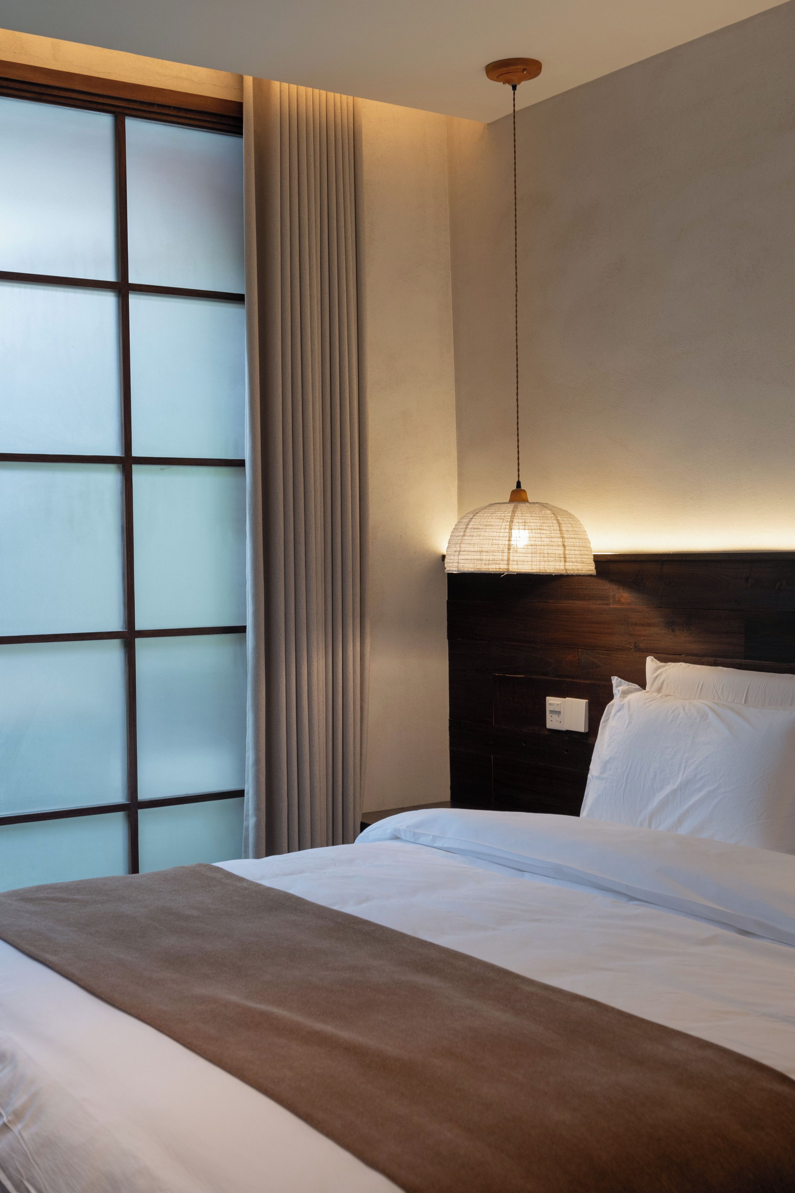 Una habitación de hotel con una cama, cabecera, lámpara colgante y ventana con cortinas.