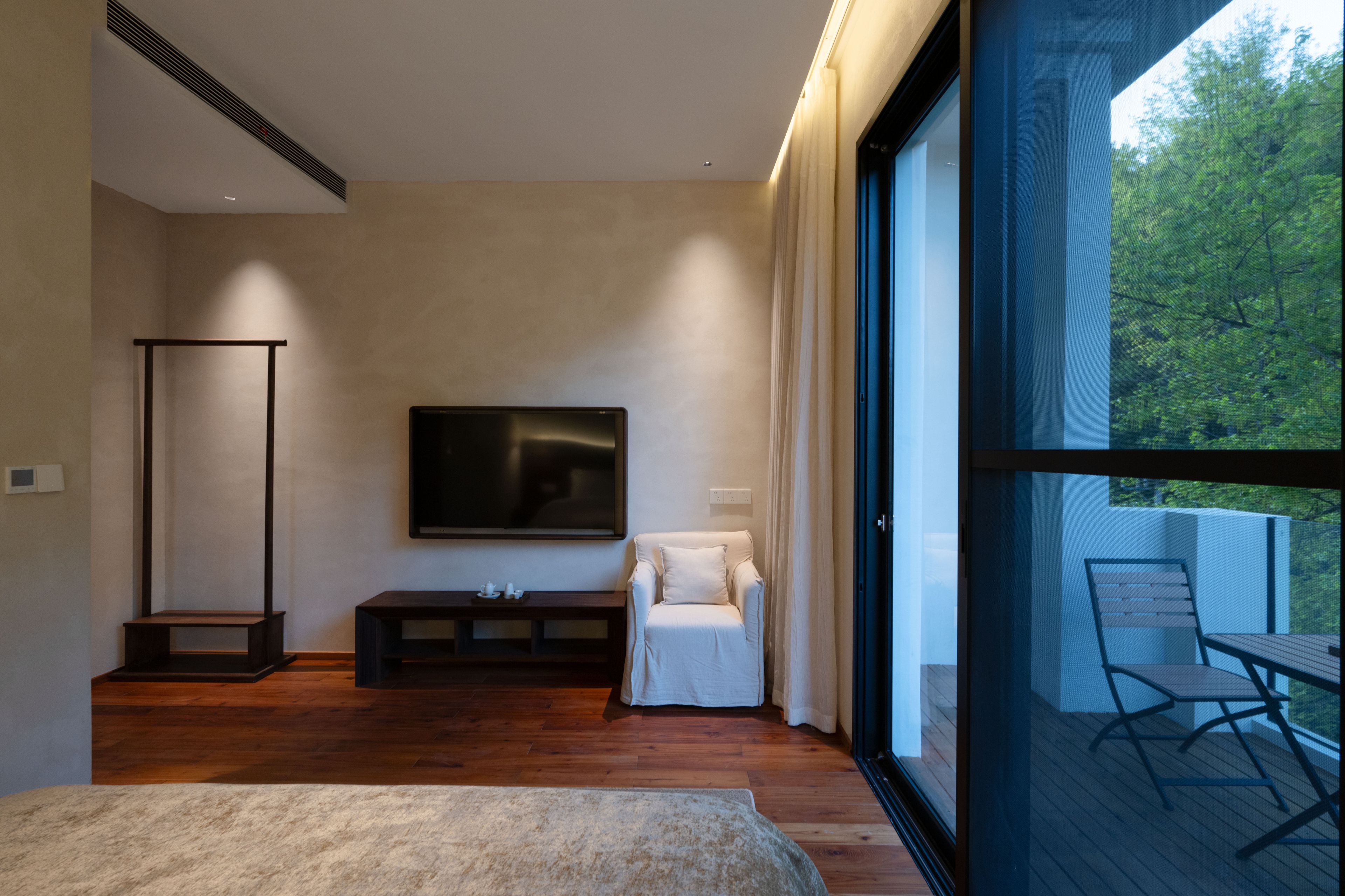 A modern bedroom with a large bed, a TV mounted on the wall, a seating area with a sofa, and a balcony with a table and chairs.