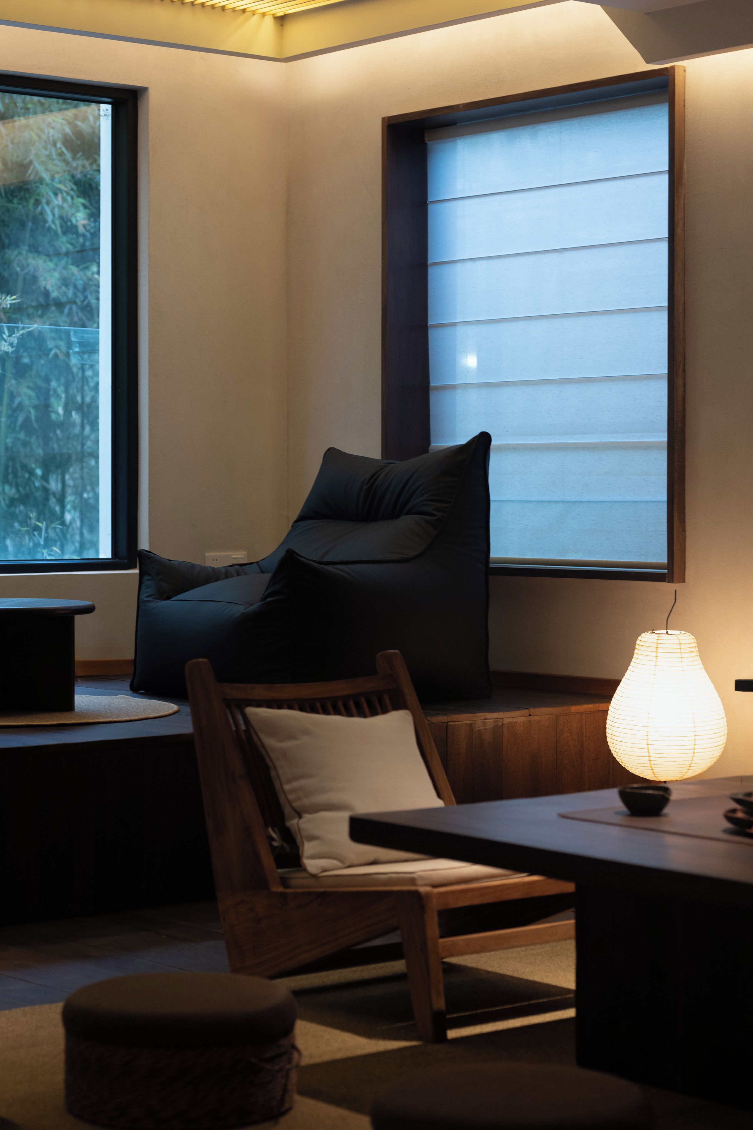 Een kamer met een houten stoel, een tafeltje, een lamp en een raam met jaloezieën.