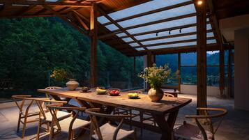 Un área de comedor al aire libre con mesa y sillas de madera, rodeada de paredes de vidrio y vista a una zona boscosa.