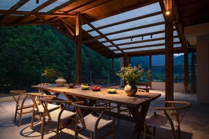 Un área de comedor al aire libre con mesa y sillas de madera, rodeada de paredes de vidrio y vista a una zona boscosa.