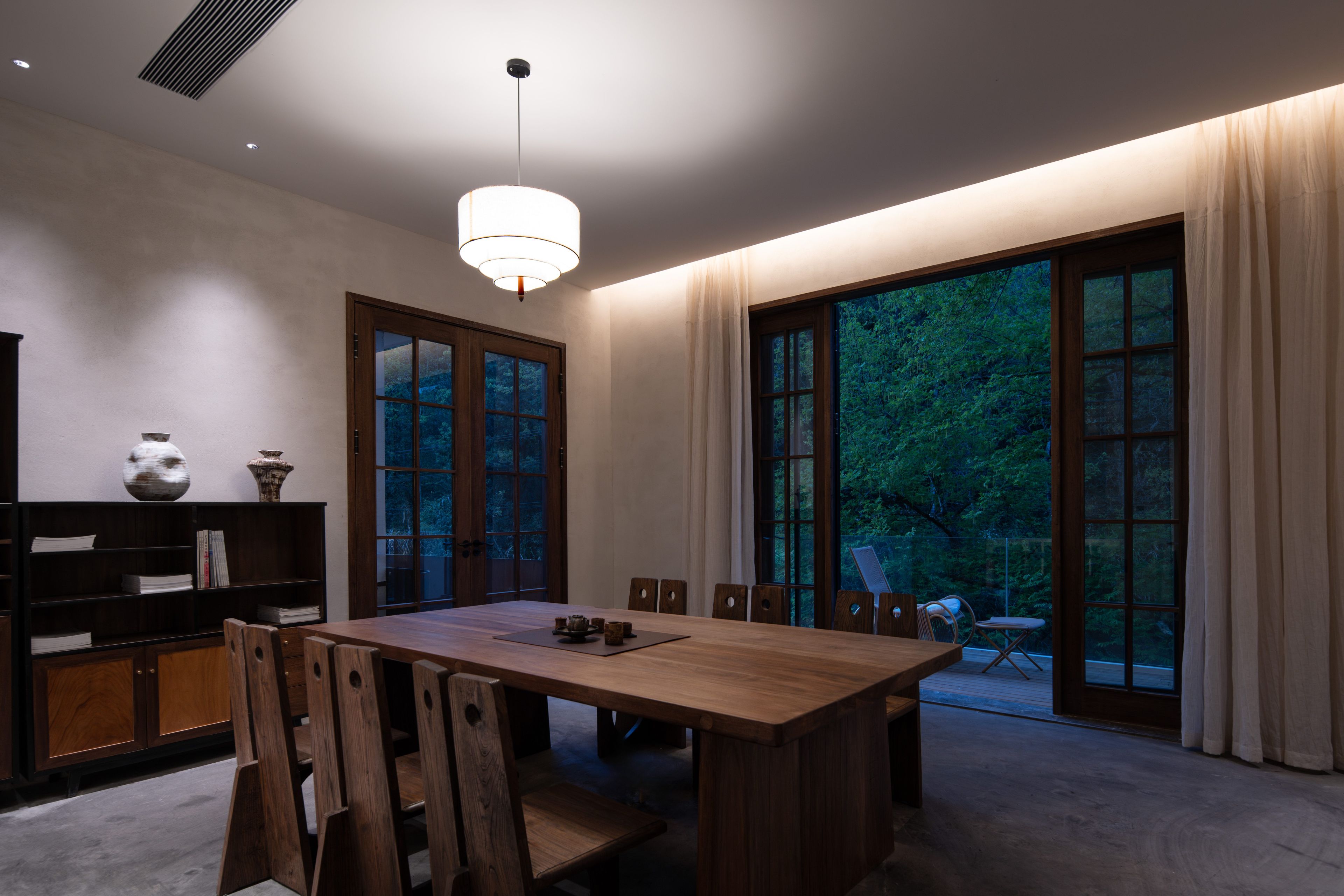Un comedor con mesa y sillas de madera, una estantería y ventanas grandes con cortinas.