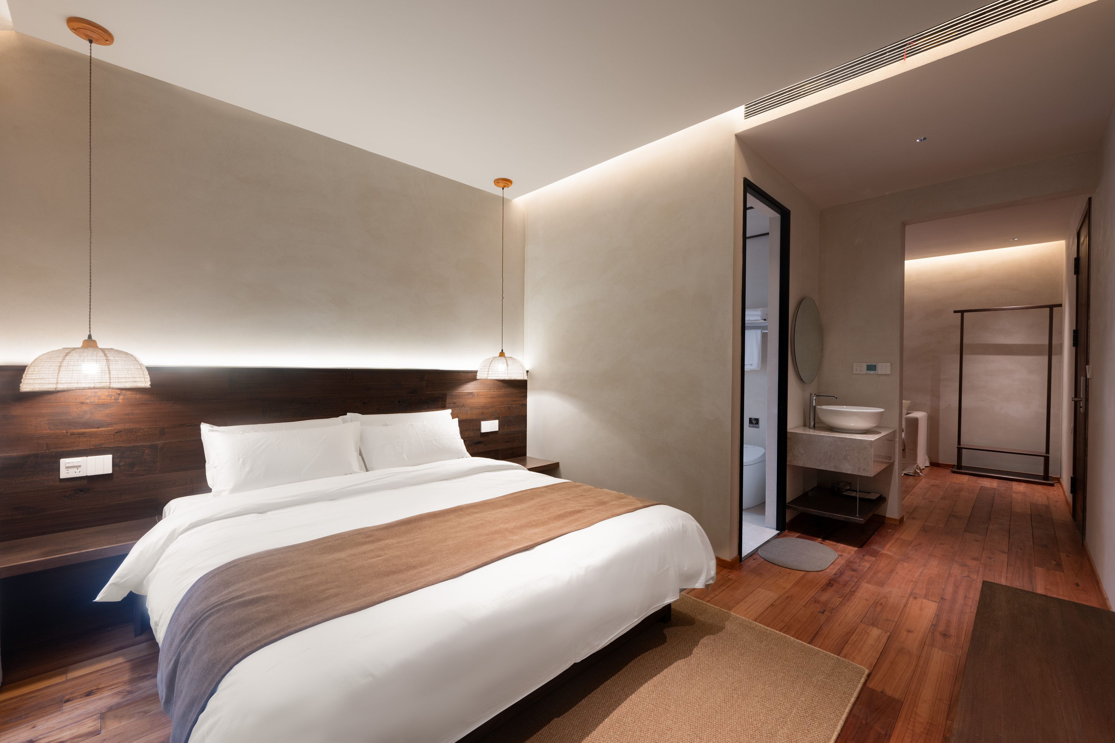A modern hotel room with a large bed, a wooden headboard, a hanging light fixture, and a bathroom with a sink and mirror.