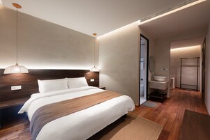 A modern hotel room with a large bed, a wooden headboard, a hanging light fixture, and a bathroom with a sink and mirror.
