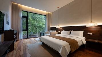 A modern bedroom with a large bed, a desk, and a view of greenery through the window.