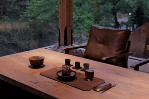 Una mesa de madera con una tetera, tazas y un ratón. Hay una silla de cuero y una vista de árboles a través de una ventana.