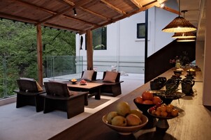 A modern outdoor dining area with a wooden table and chairs, a bowl of fruit, and a hanging light fixture.