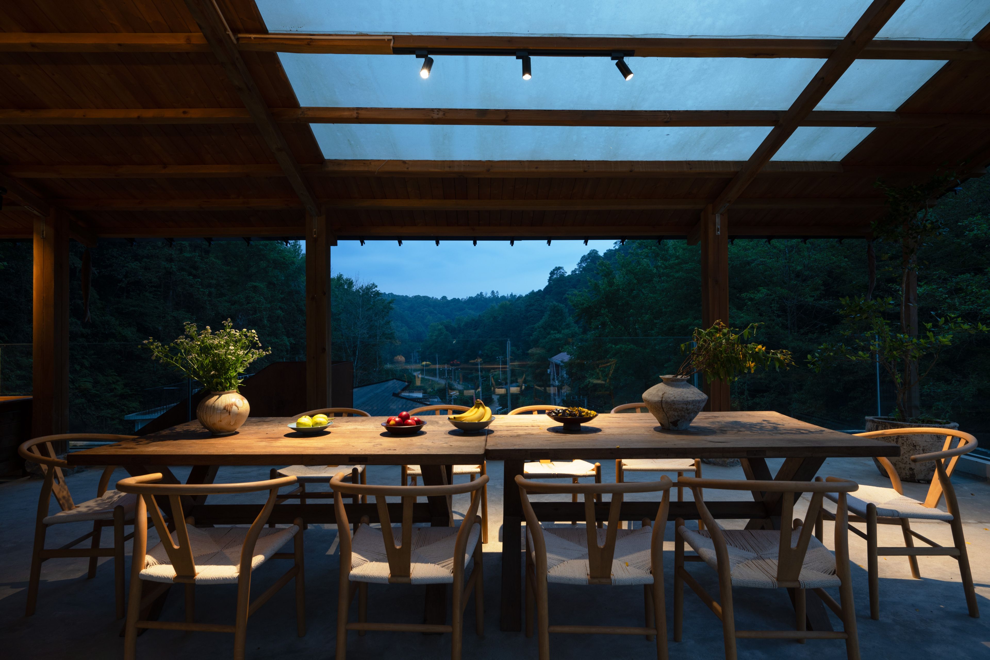 Un espace de restauration en plein air avec une table et des chaises en bois, entouré d’une pergola en bois dotée de panneaux de verre, offrant une vue sur un paysage boisé.